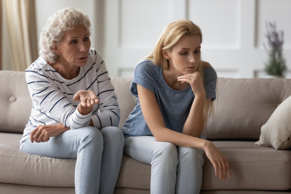 Displeased young woman sitting on sofa trying to avoid conflict with scolding older woman